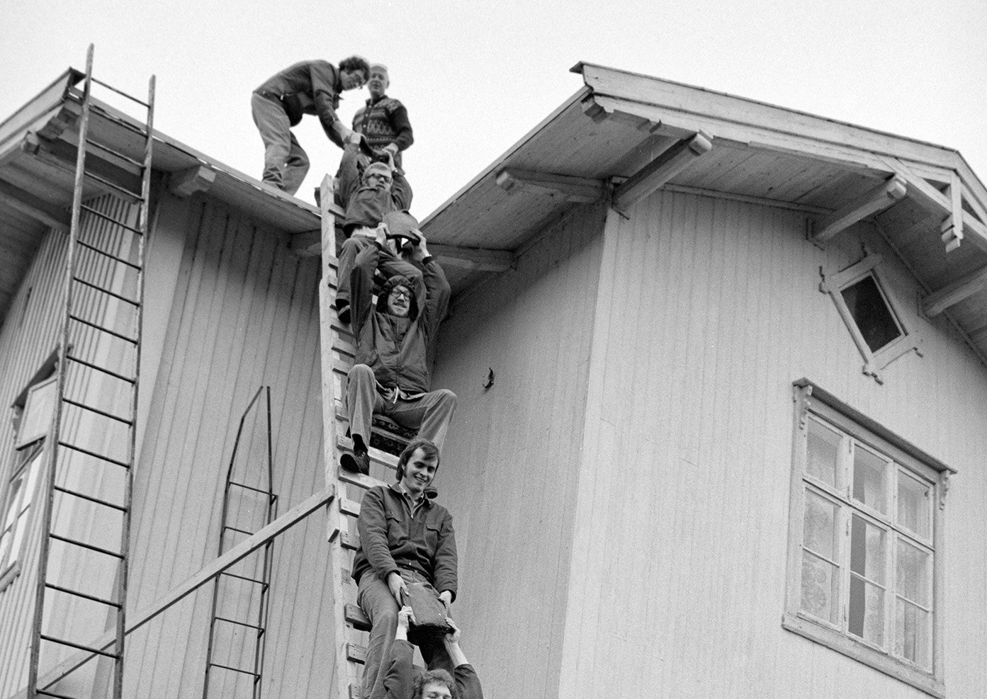 Historisk svart-hvitt-bilde av en gjeng som sitter oppetter en stige langs veggen på et hus under dugnad.
