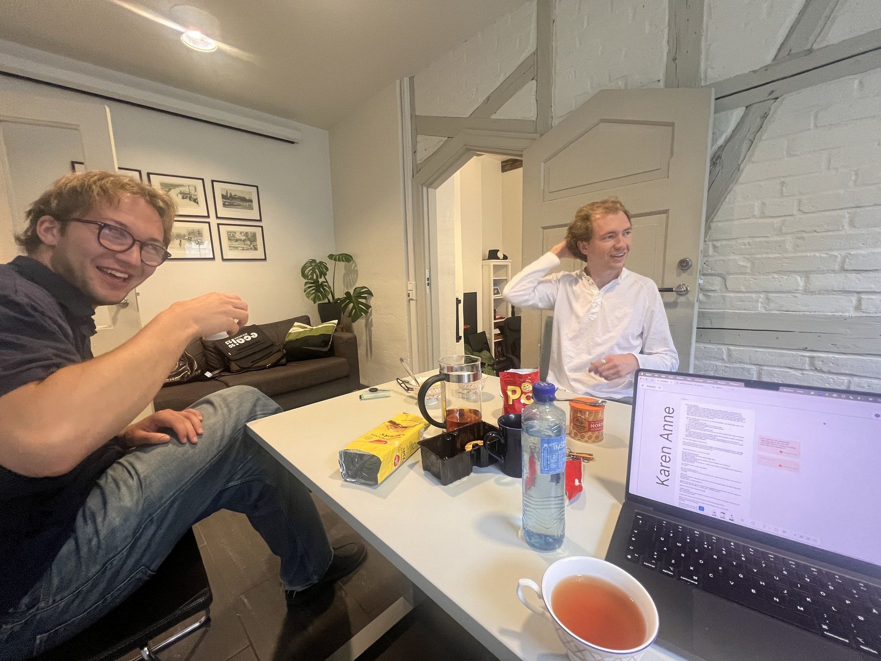 To deltakere ler rundt et bord med tekanne, kjeks og en åpen bærbar PC under den interne designworkshopen.